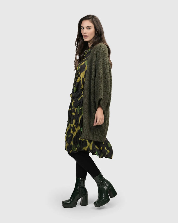 A woman wearing a cozy ALEMBIKA Sunday Cardigan, Hunter green, long knit cardigan dress and black boots.