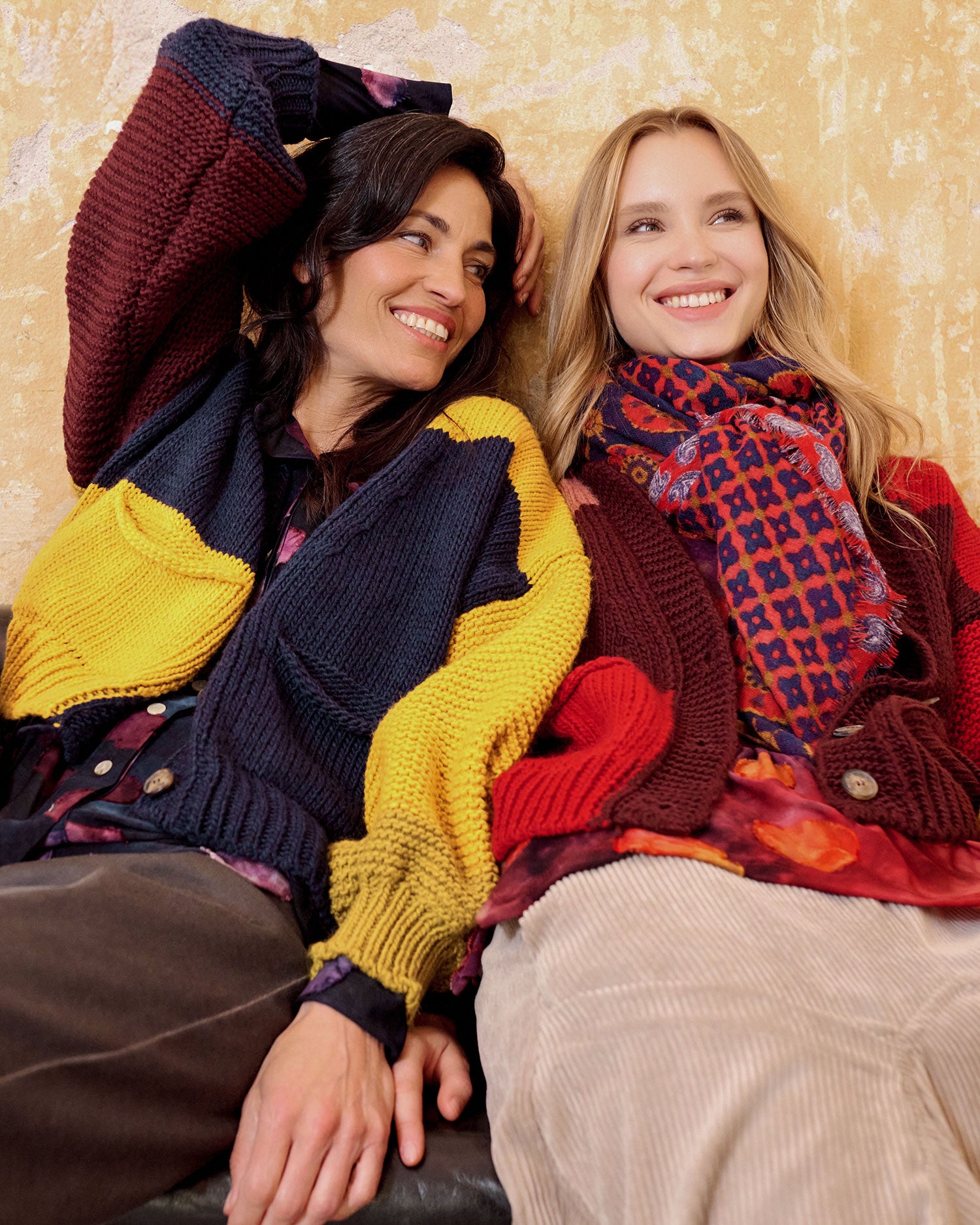 Two women smiling and posing together in cozy, colorful chunky knit sweaters and scarves against a textured beige wall.