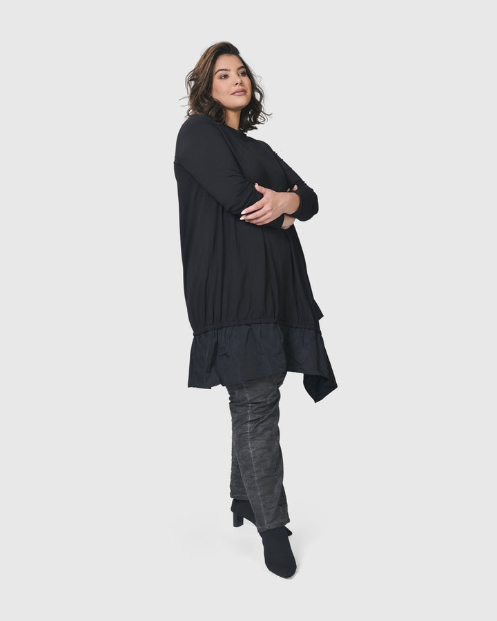 A woman wearing an ALEMBIKA Urban New Wave Tunic Top in Black with long sleeves and grey pants.