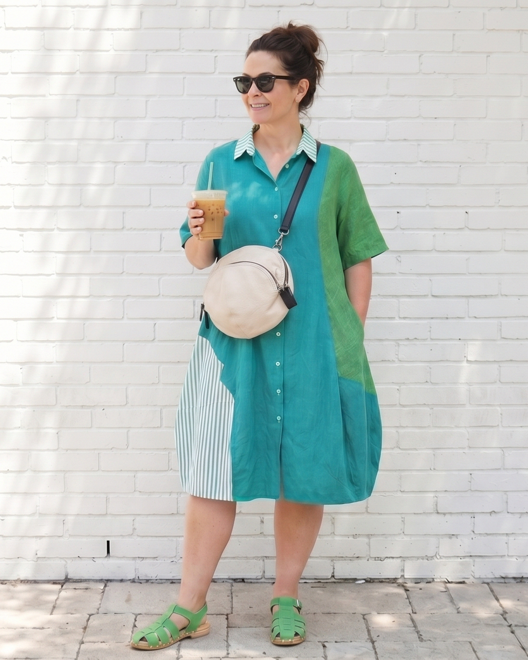 Woman wearing a green and teal dress holding a drink against a white brick wall.