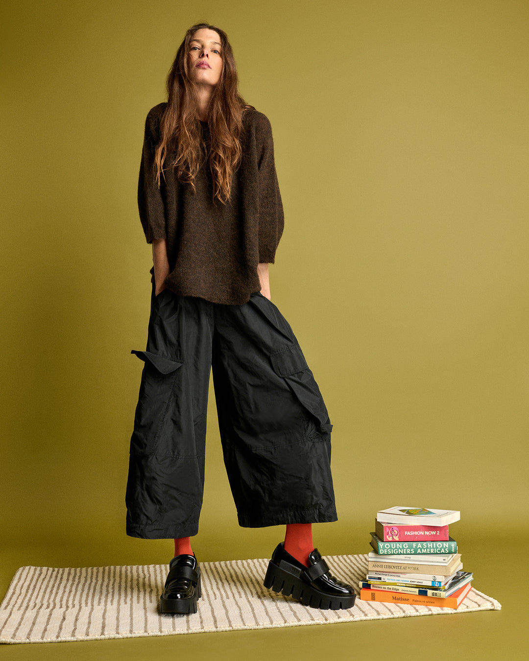 Woman in brown top and black wide-leg pants standing on a green background with books.