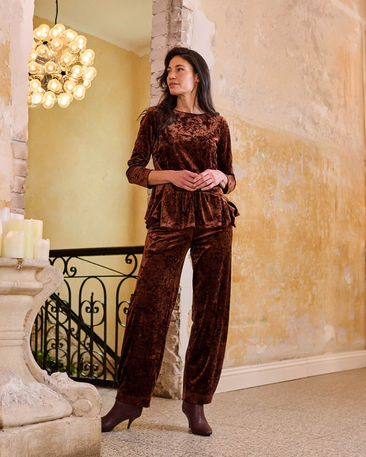 A woman models a matching brown velvet lounge set consisting of a long-sleeve top and wide-leg pants, standing in a warm, rustic interior with a chandelier and textured walls.
